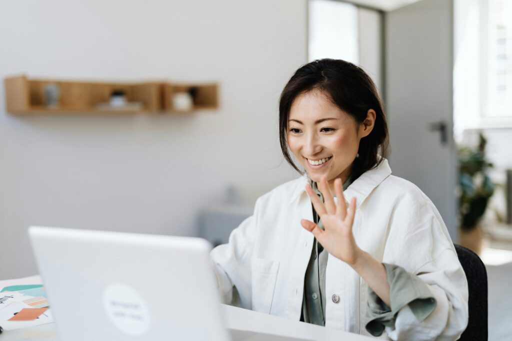 Smiling woman waves at laptop during a video call in a modern office.